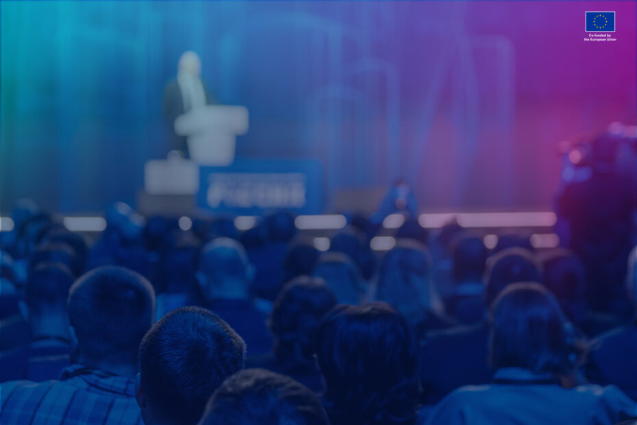 Blurred audience listening to a speaker at a professional conference event in a large auditorium.