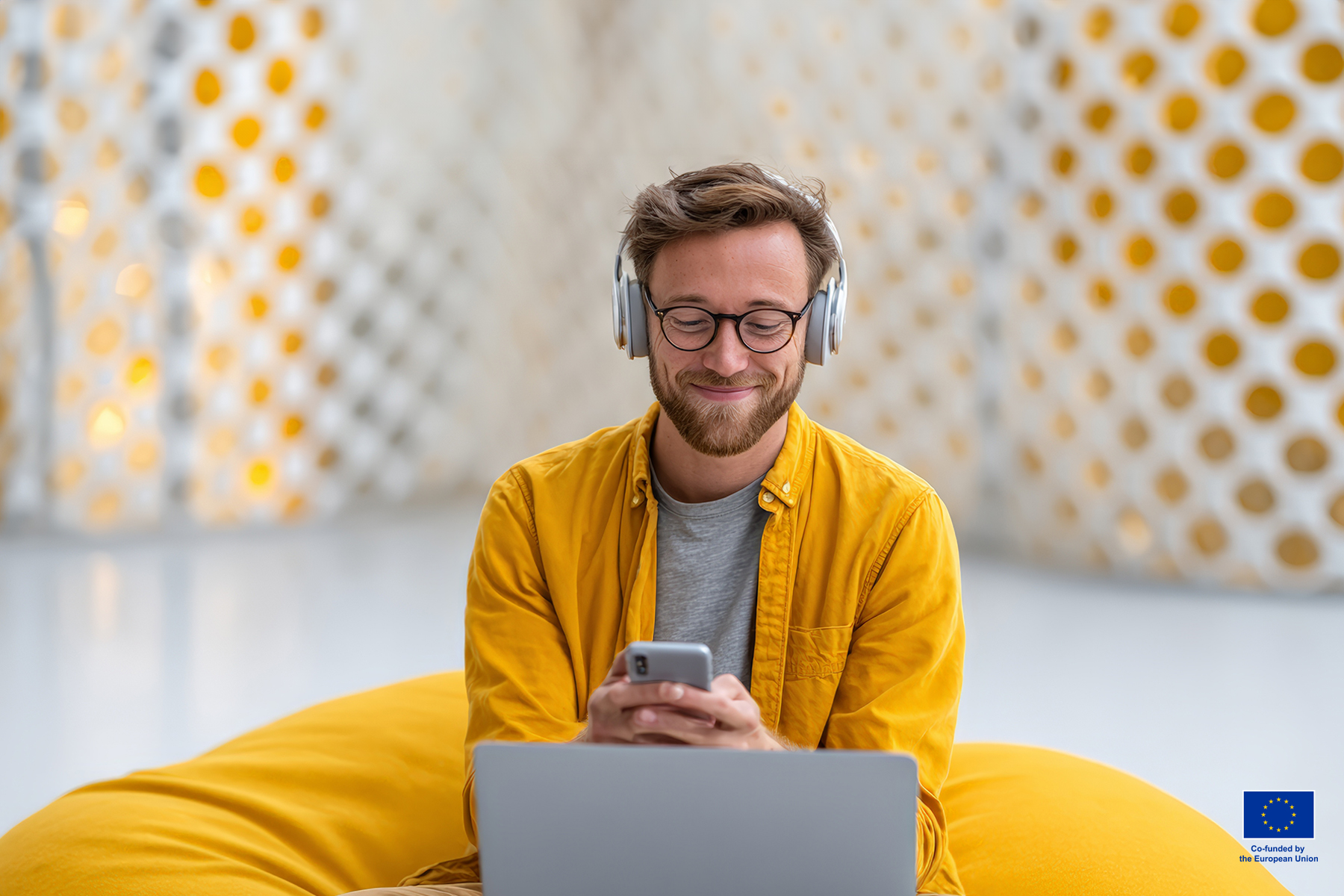 Gen Z man sitting front of a lap top and smiling. He is holding a mobile phone and watching it. He is wearing headphones.