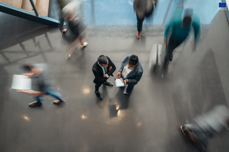 Busy office, two people standing in the middle as others are blurred because of the shutter speed usage.