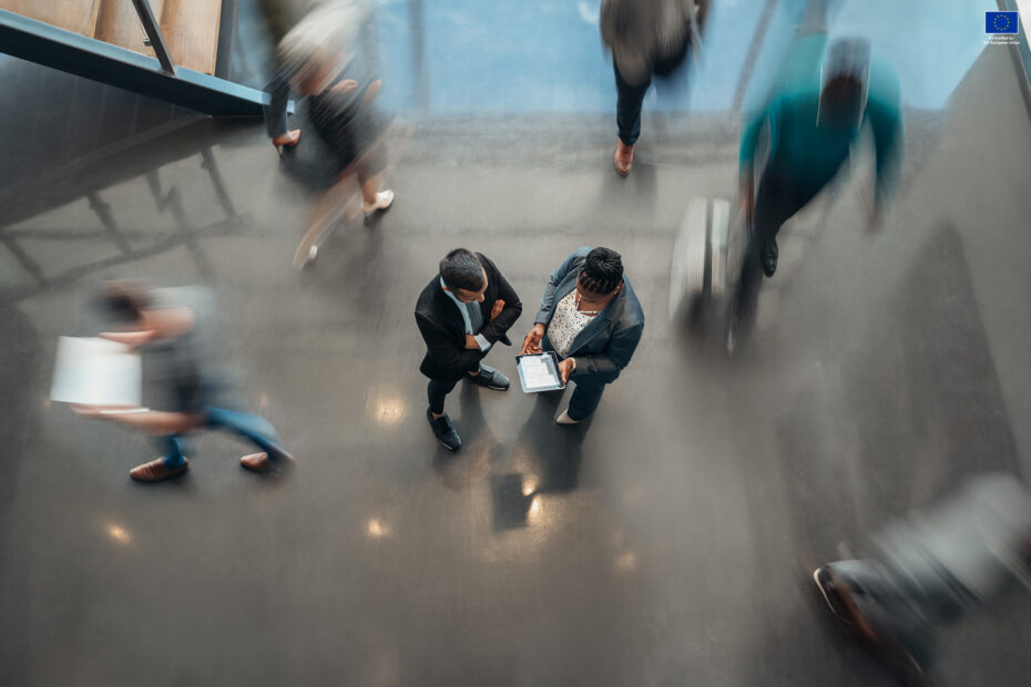 Busy office, two people standing in the middle as others are blurred because of the shutter speed usage.