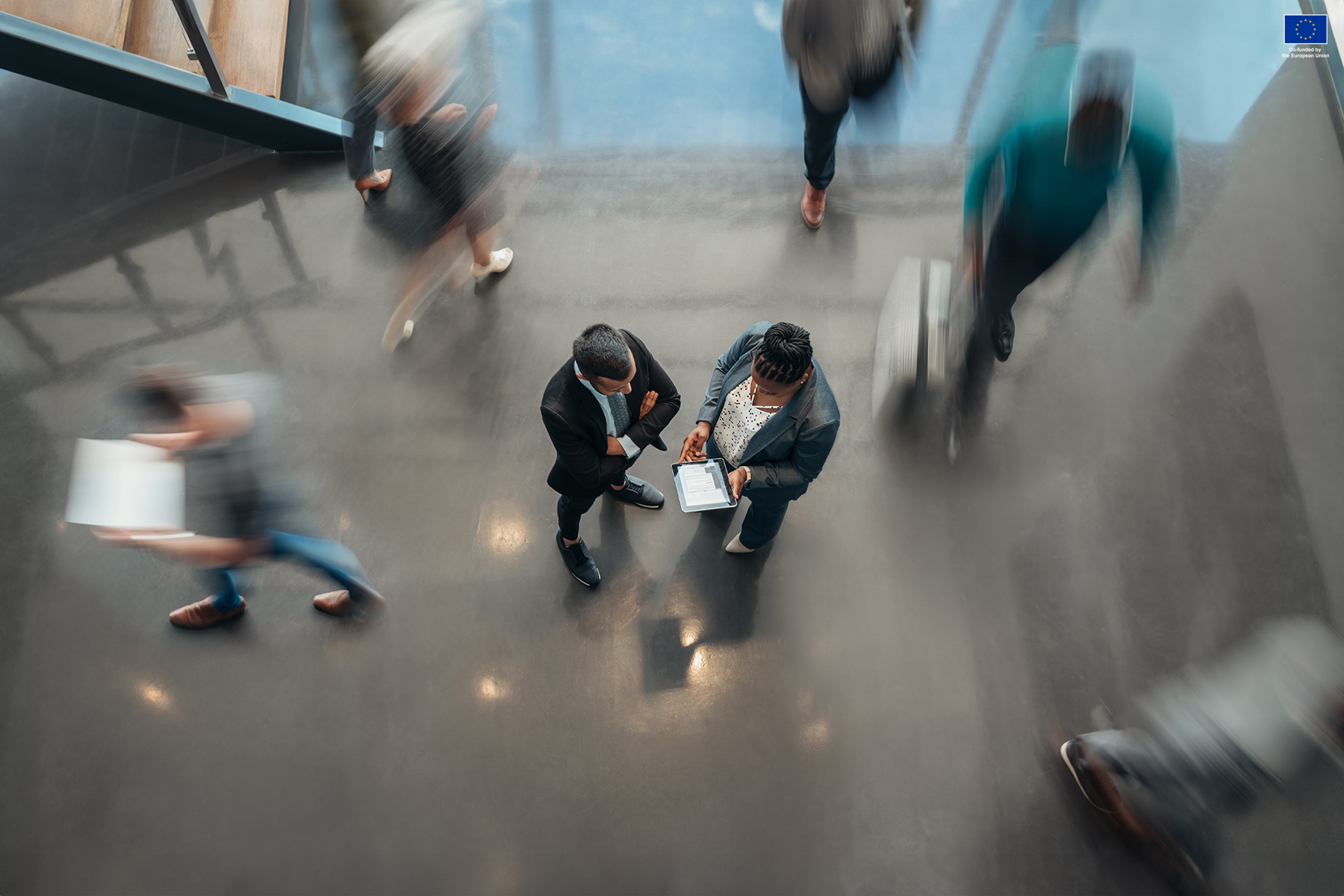 Busy office, two people standing in the middle as others are blurred because of the shutter speed usage.
